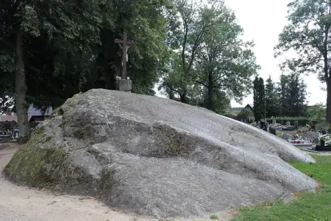 Ogromny głaz narzutowy na cmentarzu, zwieńczony krzyżem. Otoczony drzewami i nagrobkami.
