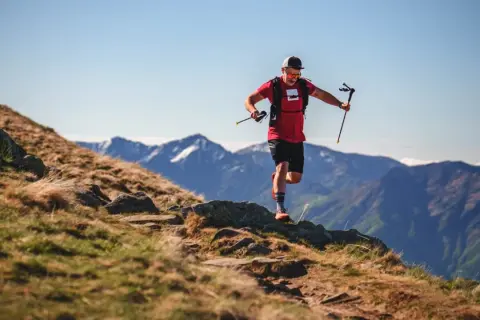 Ein Mann im roten Shirt und schwarzer Hose läuft mit Stöcken einen Bergpfad entlang. Sein intensives anaerobes Training fordert ihn in der atemberaubenden Berglandschaft.
