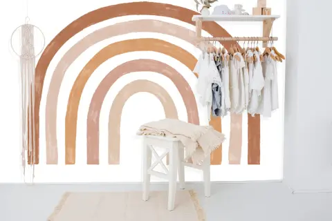 Un arc-en-ciel aux tons terreux est peint sur un mur blanc, orné d'une penderie d'enfants et d'une décoration murale en macramé.
