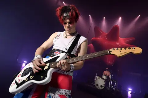 Yungblud gra na gitarze podczas koncertu w Warszawie. W tle czerwona, dmuchana instalacja i perkusista.