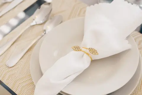 Une serviette blanche mariage, ornée d'un anneau doré scintillant, repose sur une assiette. Des couverts argentés sont disposés à côté.