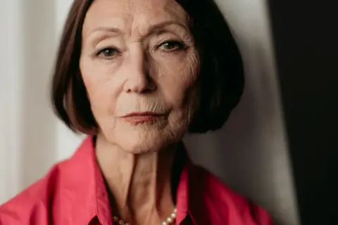 Portret starszej kobiety, Ewy Woydyłło, z widocznymi zmarszczkami, w różowej bluzce i perłach. Jej życie prywatne jest pełne doświadczeń.