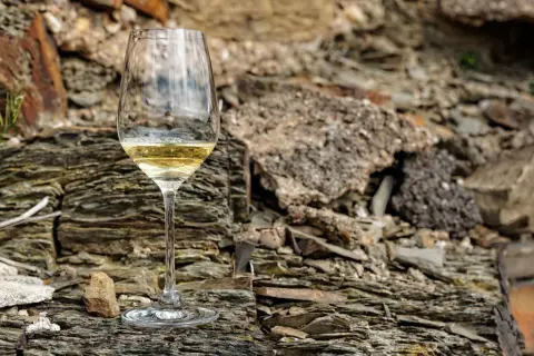 Ein Glas Weißwein steht auf schieferigem **Weinboden**. Die raue Gesteinslandschaft bildet einen starken Kontrast zum edlen Tropfen.