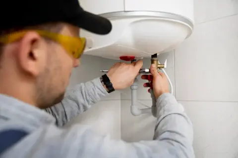 Instalator w okularach ochronnych pokazuje, jak podłączyć bojler elektryczny, pracując przy zaworze.