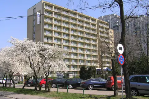 Hotel Felix Warszawa w otoczeniu kwitnących drzew. Widać parking z samochodami i budynek hotelu z balkonami.