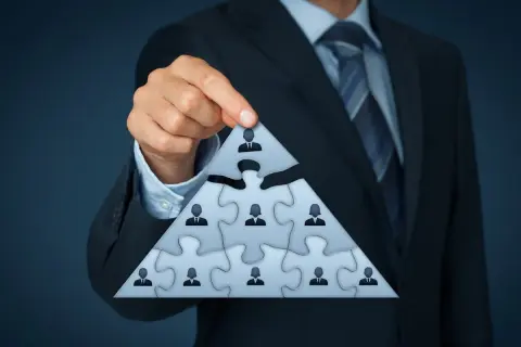 Biznesmen układa piramidę z puzzli, symbolizującą strukturę firmy. Każdy element to pracownik, a najwyższy to beneficjent, który czerpie korzyści.
