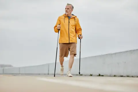 Mężczyzna w żółtej kurtce i brązowych spodenkach wykonuje rozgrzewkę przed nordic walking, trzymając kije.