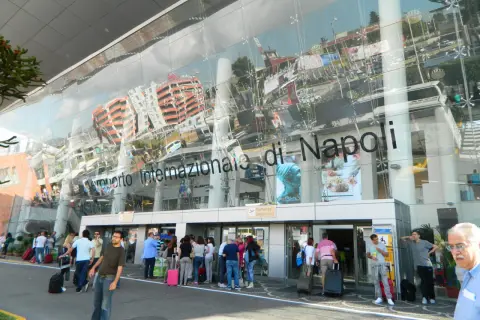 Ludzie z bagażami przed nowoczesnym budynkiem Aeroporto Internazionale di Napoli.
