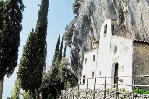 Piccolo eremo di San Valentino incastonato nella roccia, circondato da alti cipressi e un cielo azzurro.