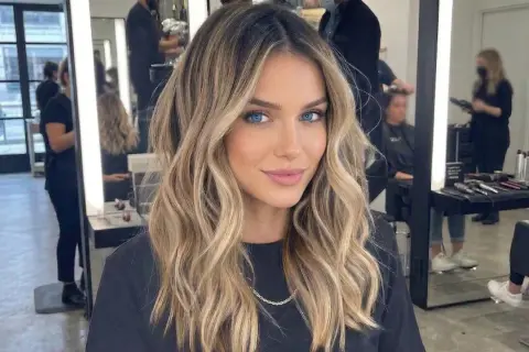 Kobieta z pięknymi, falowanymi blond włosami, z subtelnym, ciemnym odrostem przy blondzie. W tle salon fryzjerski.