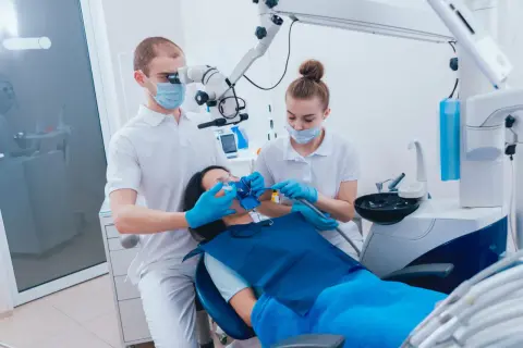 Dentysta pod mikroskopem wykonuje ekstyrpację miazgi u pacjentki, asystuje mu pomoc dentystyczna.
