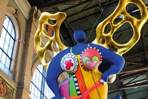 Silhouette d'une Nana colorée de Niki de Saint Phalle, avec des ailes dorées, suspendue dans un grand hall.