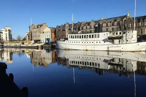 Barcos y edificios históricos reflejados en el agua en Leith, Edimburgo. El "Ristorante De Niro" en un barco blanco destaca.