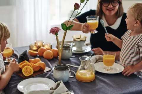 Familjen samlas kring frukostbordet med apelsinjuice och frukt. Duken är av linne, redo att stryka för nästa måltid.