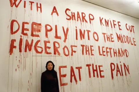 A woman stands before a wall with red, dripping text that reads "WITH A SHARP KNIFE CUT DEEPLY INTO THE MIDDLE FINGER OF THE LEFTHAND EAT THE PAIN." This is a reference to "spirit cooking.