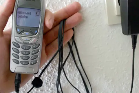 Dłoń trzyma starą Nokię, która ładuje się przez kabel. Na ekranie telefonu widnieje napis "Akku wird geladen". To klasyczny przykład technologii 3G z dawnych lat.