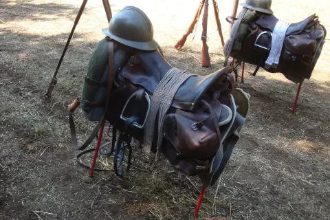 Dwa siodła, część rzędu końskiego, z hełmami i karabinami w tle.