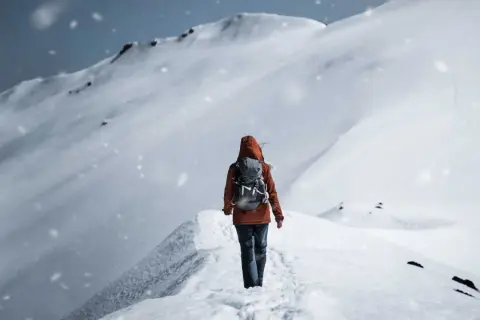 Persona con zaino cammina su un crinale innevato, mentre fiocchi di neve cadono dolcemente.