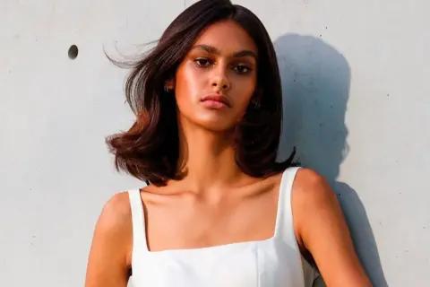 Mujer con cabello castaño brillante y piel bronceada, luciendo un vestido blanco. Su cabello parece suave, como si hubiera aprendido como se usa el acondicionador.