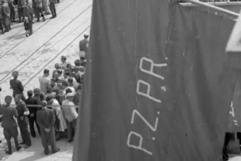 Tłum ludzi zgromadzony na ulicy, na pierwszym planie widoczna wielka, ciemna tkanina z napisem PZPR.