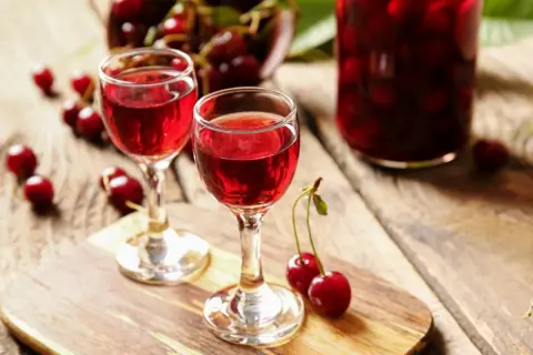 Dwa kieliszki z rubinowym winem z czereśni, obok świeże owoce i duży słój z nalewką.