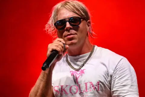 Artysta Skolim w okularach i białej koszulce z napisem "SKOLIM" na czerwonym tle. Zastanawiasz się, ile Skolim bierze za koncert?