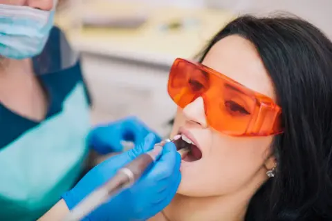 Pacjentka w okularach ochronnych przechodzi zabieg znieczulenia komputerowego zęba. Dentysta w rękawiczkach używa nowoczesnego urządzenia.