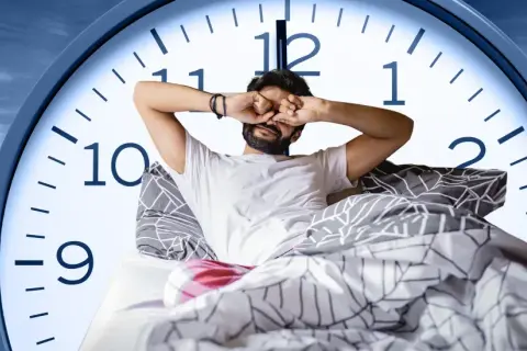 Man in bed rubs his eyes, looking tired. A large clock face behind him shows it's just past 6 AM, suggesting only six hours of sleep.