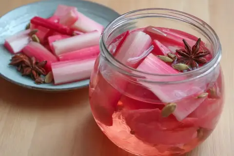 Rabarbersaft recept: färska rabarberbitar i en glasburk med kryddor som stjärnanis och kardemumma. En tallrik med rabarber och kryddor står bredvid.
