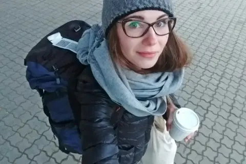 Kobieta w czapce i szaliku trzyma kubek kawy, obok plecak.