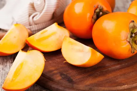 Saftige, leuchtend orangefarbene Persimmon-Früchte, einige ganz, andere in Spalten geschnitten, liegen auf einem Holzbrett.