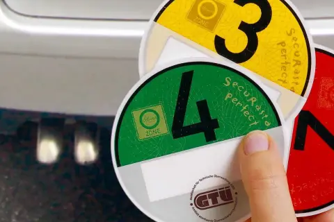 Umweltplaketten für Umweltzonen in Europa. Eine Hand hält grüne und gelbe Plaketten mit den Zahlen 4 und 3.