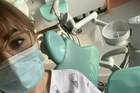 Praktyki asystentka stomatologiczna w gabinecie. Widoczny fragment twarzy w maseczce, okulary i fartuch z motywem zębów.