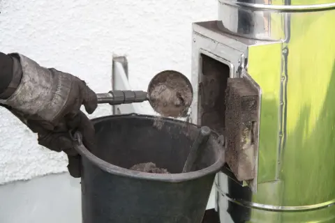 Reinigung des Bullerjan-Ofens. Asche wird mit einer Schaufel in einen Eimer gefüllt. Ein Feinstaubfilter ist hier nicht zu sehen, aber die Reinigung ist ein Schritt zur Wartung.