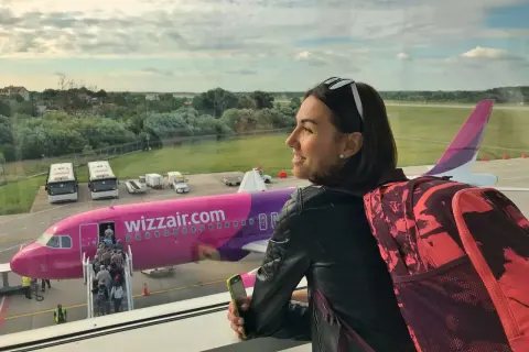 Kobieta z plecakiem i telefonem czeka na lot Wizzair. W tle różowy samolot i pasażerowie wsiadający na pokład.