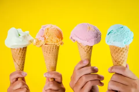 Vier Hände halten bunte Speiseeis-Sorten in Waffeln vor gelbem Hintergrund.