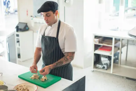 Chef con gorra y delantal a rayas, cortando champiñones en una tabla verde. Ideal para aprender a cocinar.