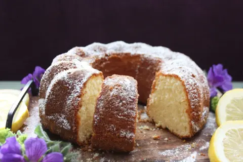 En saftig citron sockerkaka, pudrad med florsocker, serveras med citronskivor och lila blommor på en träbricka.