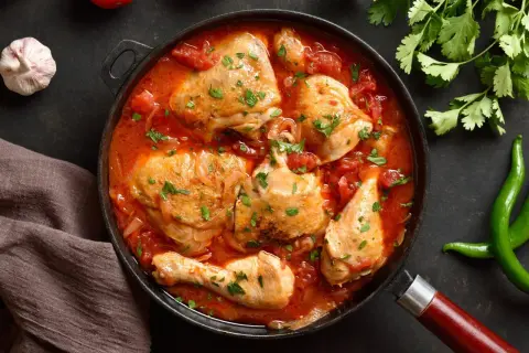 Pyszny kurczak w sosie pomidorowym, duszony z cebulą i posypany natką pietruszki. Obok czosnek, pomidory i papryczki chili.