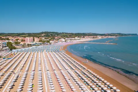 Fano mare: file ordinate di ombrelloni bianchi e colorati sulla sabbia dorata, con il mare azzurro e la costa sullo sfondo.