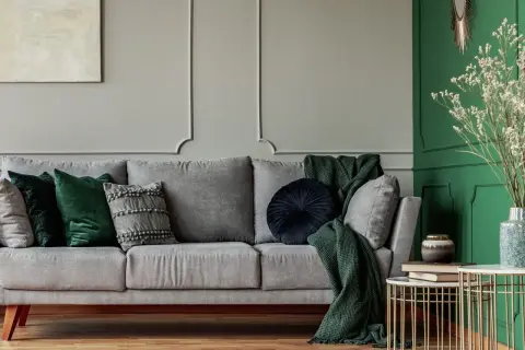 Elegancki salon w odcieniach butelkowej zieleni i szarości. Szara kanapa z zielonymi i szarymi poduszkami, zielony pled.