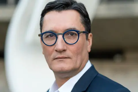 Olivier Dartigolles, un homme aux lunettes bleues, pose pour un portrait. Il est vêtu d'un costume sombre, évoquant une image de famille et de professionnalisme.