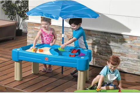 Drei Kinder genießen Sommerspiele im Sand- und Wasserspieltisch unter einem blauen Sonnenschirm.