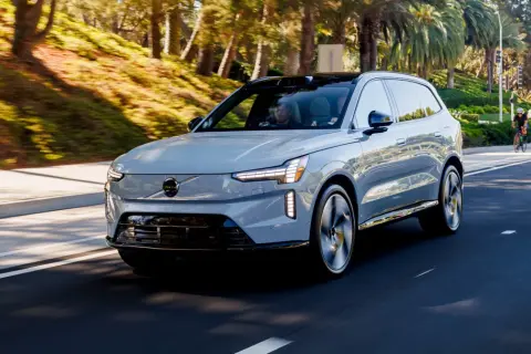 Szary Volvo EX90 jedzie po słonecznej drodze, otoczony zielenią i palmami.