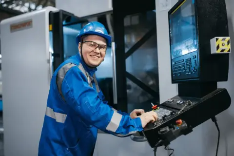 Uśmiechnięty pracownik w niebieskim kombinezonie i kasku obsługuje maszynę CNC. Zastanawia się, ilu pracowników może mieć brygadzista.