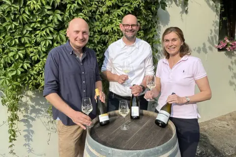 Drei Personen mit Wein vor einer Weinrebenwand. Die Flasche ganz rechts trägt das Etikett "Auxerrois".