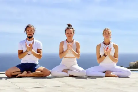 Tres personas meditan al aire libre, con las manos juntas en un mudra. El mar y el cielo azul forman el telón de fondo.
