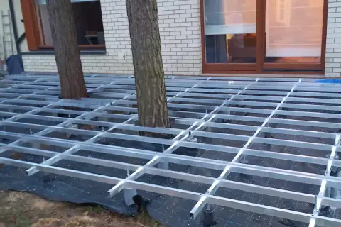 Trwa układanie deski kompozytowej na aluminiowym stelażu, który omija drzewa rosnące na tarasie.
