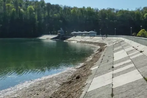 Zapora Myczkowce z zieloną wodą i betonowym zboczem. W tle las i budynki techniczne.