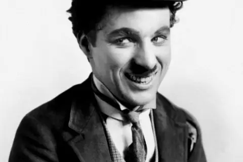 Charlie Chaplin w meloniku i garniturze, z uśmiechem, jakby opowiadał dowcip swoim dzieciom.